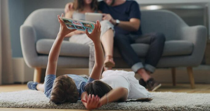Carefree young boy and girl lying on the floor in the lounge and having fun using a modern digital tablet together. Brother and sister relaxing online with their parents in the background at home