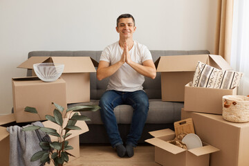 Portrait of man wearing white T-shirt and jeans sitting on sofa surrounded with pile of packages,...