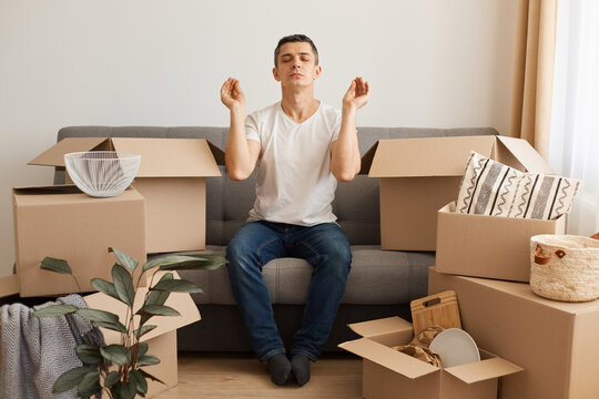 Indoor shot of man wearing white T-shirt and jeans sitting on cough surrounded with pile of packages, trying to relax via yoga on day of move to rented home, unbox carton box with belongings.