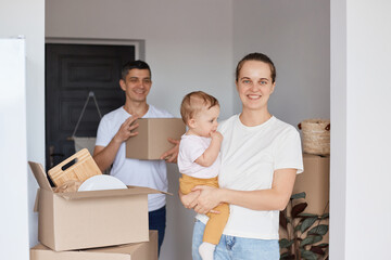 Fototapeta na wymiar Portrait of happy family with little daughter posing in their new apartment surrounded with boxes with belongings, looking at camera with positive facial expressions, relocation and delivery service.