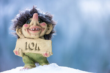 Fototapeta na wymiar Cute happy troll holding sign with the word 10k chiseled out. Soft blurred out background in winter wonderland.