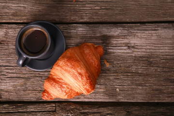 Expresso cup and croissant on old rustic wooden table background. Fresh baked bun and hot black coffee top view. Food, French breakfast, good morning, cafe menu concept. Close up, copy space
