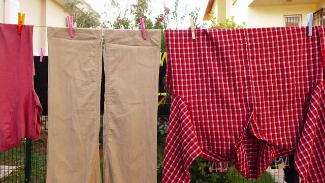 Beige pants and red checkered shirt are hanging on the clothesline attached by pins after laundry outdoors. Linen textile is drying and trembled by wind after being washed on the backyard of the house
