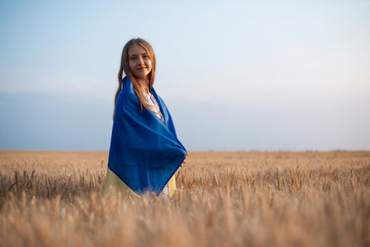 Smiling girl wrapped in ukrainian flag in the boundless field of rye at sunset. Patriotic ukrainian girl