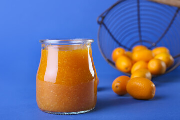 Jar of tasty kumquat jam and fresh fruits on blue background