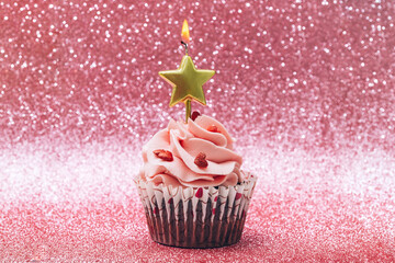 Cupcake with burning star shaped candle on glitter background. Happy birthday concept.