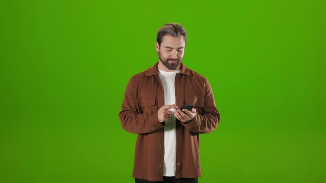 Caucasian bearded man standing over chroma key background with modern smartphone in hands. Casually dressed guy surfing internet in studio.
