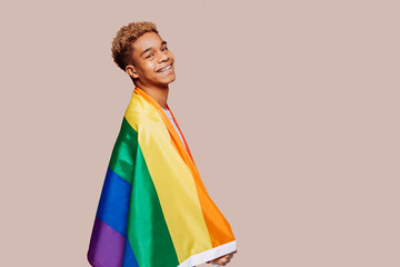 Side view portrait of cheerful young afro latin black man with a gay pride rainbow flag at studio over beige background. Homosexual lgbtiq concept, rainbow flag, celebrating parade. Copy space. - Powered by Adobe