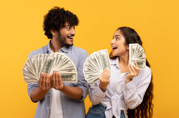Emotional indian couple holding bunch of money banknotes and looking at each other, yellow...