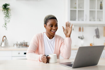 Cheerful young pretty black woman with cup of favorite drink, waves hand to laptop webcam