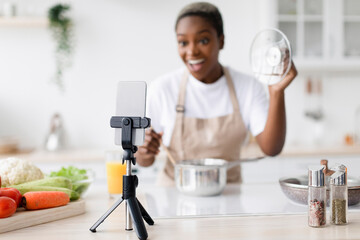 Smiling surprised millennial black female with open mouth in apron prepares lunch shoots video for blog on smartphone