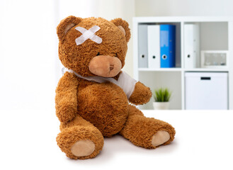 medicine, healthcare and childhood concept - teddy bear toy with bandaged paw and patch on head over medical office at hospital background