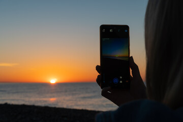 Close-up of women's hands with a mobile phone that vertically photograph a beautiful sunset on the...