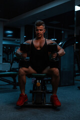 Fototapeta na wymiar Pretty fashionable young man bodybuilder with muscular body sits and does dumbbell exercises in the gym on a dark background