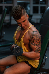 young sports man bodybuilder with a maya tattoo sits and pumping his biceps in the gym.