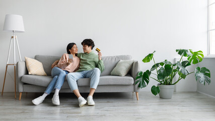 Naklejka premium Cheerful young Asian couple sitting on sofa with credit card and cellphone, shopping online in living room, copy space