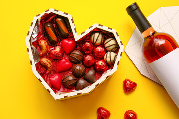 Heart shaped box with tasty chocolate candies and bottle of wine on yellow background