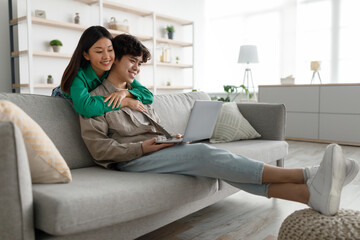 Young Asian woman hugging her boyfriend, using laptop, browsing web on couch at home, empty space