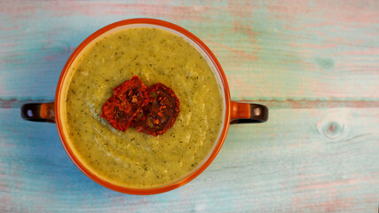 on a blue wooden table there is a brown plate with green broccoli soup with dried tomatoes on top. top view. gluten and lactose free