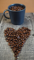 a dark gray mug filled with roasted whole coffee beans stands on a wooden background with coffee beans laid out in the shape of a heart. side view