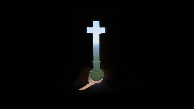 Crosslet loop or embrasure in wall of old castle, green landscape seen through narrow opening. Firing slot in shape of inverted keyhole with cross, old fortification on top of hill