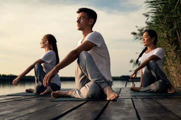 Group of people doing yoga exercises by the lake at sunset.