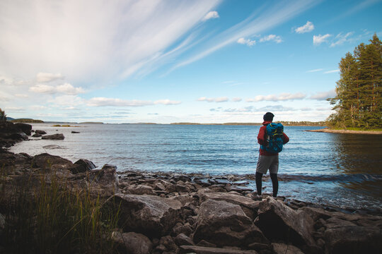 Hiker wearing a jacket and carrying a backpack walks along the beach and watches Lake Jatkonjarvi at sunset in Koli National Park, eastern Finland. Man aged 24 wearing sports clothes. Active lifestyle