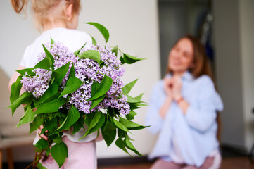 Mothers Day. A little preschool girl holds a bouquet of lilac flowers behind her back and wants to give it to her mom