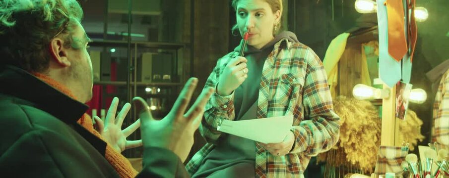 Young female scriptwriter holding paper and marker pen while discussing circus act with clown in dressing room