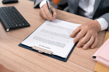 A document, an employment contract signed by the employer, boss, manager, hands of an elderly man signing on a piece of paper, sitting at a desk across from a female employee