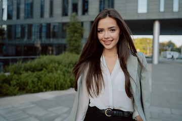 Smiling girl with brown hair walks out of corporate building for break dressed in smart shirt and jacket thrown over back, walks outside past glass skyscraper, secretary, businesswoman, boss