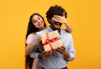 Pretty indian woman covering her boyfriend's eyes, holding gift box and greeting him with birthday...