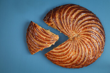 Top view of traditional French galette des rois (king cake) with cut piece, blue background. Cake made with puff pastry and creamy almond filling roll in circles shape. It's usually served on Epiphany
