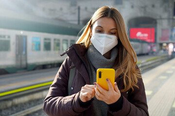 Portrait of a girl in a protective mask standing on the platform of train station with a mobile...