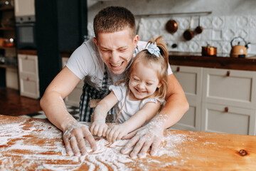Cheerful dad and daughter child in aprons cook together have fun playing with flour sitting at a wooden table in a cozy kitchen at home. Selective focus