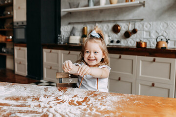 A cheerful happy emotional little girl in an apron cooks, laughs and plays with flour sitting at a wooden dining table in a cozy kitchen at home. Selective focus