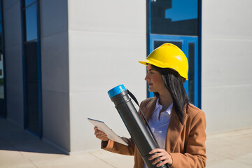 Young beautiful woman in brown coat, yellow workman's helmet and blueprint tube, working with her tablet. Business concept, architect, construction, working woman, empowerment.