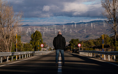 Rear view of adult man standing on asphalt road looking at view of modern windmills. Almeria, Spain