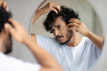 Haircare treatment. Worried indian guy looking at flaky scalp in mirror, examining gray hair and hairloss issue