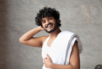 Male beauty care. Happy indian guy with towel on shoulder standing in bathroom and smiling at camera