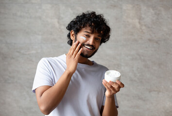 Cheerful indian man applying cream on face, holding moisturizer jar in hand, smiling to camera,...