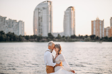 Fototapeta na wymiar Senior newlyweds hug. Stylish couple of elderly newlyweds stand embracing on river bank.