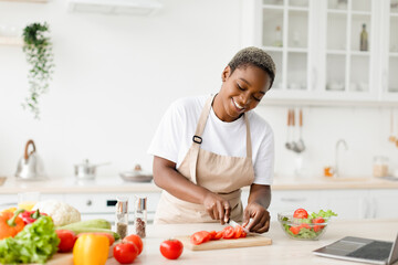 Smiling millennial pretty black female in apron prepare healthy lunch from organic vegetables, cuts...