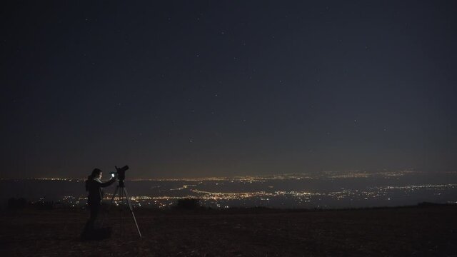 Woman photographing stars with a camera and a tripod at night. City lights in the background. 