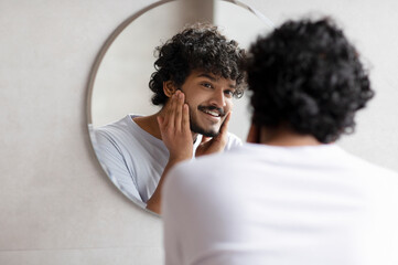 Attractive bearded indian man touching face applying moisturizer on face, standing near mirror in...