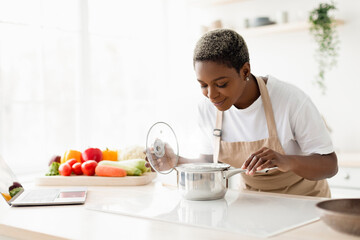 Cheerful young pretty black female in apron prepare lunch and smell dish in minimalist kitchen interior