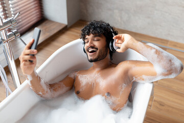 Excited indian man enjoying music in wireless headphones, relaxing in foamy bath and listening...