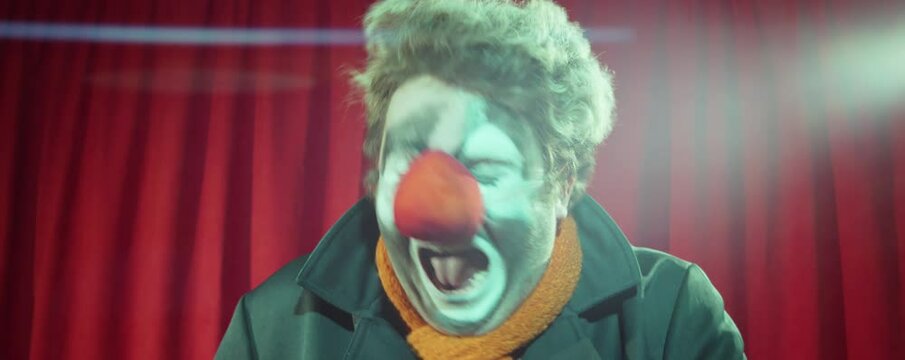 Clown standing in front of red curtain and talking in microphone while giving stand up on stage
