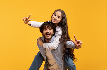 Romantic indian couple having fun together, showing V-sign gesture, guy piggybacking his...
