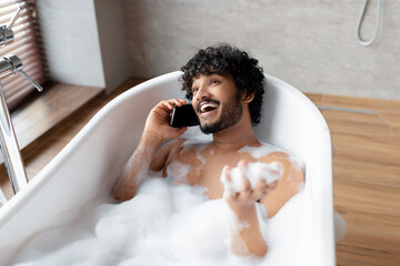 Young restful indian man talking on smartphone while lying in bathtub filled with water and foam in...